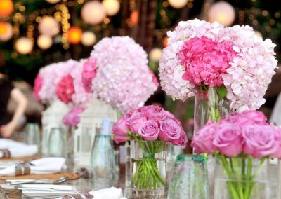 A decorated table setting that appears to be part of a larger event, possibly a wedding or banquet. There are large floral arrangements with pink blooms, some appearing to be hydrangeas and others roses, set in clear glass vases of varying heights.