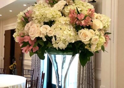 A large floral arrangement in a clear glass vase placed on a table. The arrangement consists of various flowers, including white hydrangeas, pink and white roses, and other greenery.
