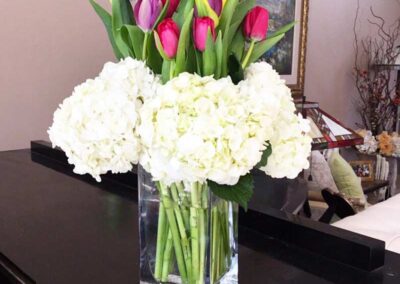 A bouquet of flowers in a clear glass vase placed on a dark surface, possibly a table. The bouquet is composed of white hydrangeas and tulips in various colors including purple, pink, and yellow.