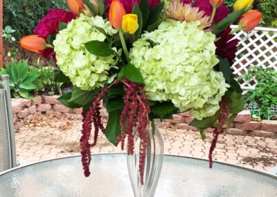 A vibrant floral arrangement in a clear glass vase. The bouquet consists of large green hydrangeas, bright red and purple flowers, yellow and orange tulips, and some hanging burgundy amaranthus
