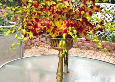 A vibrant floral arrangement featuring red and yellow flowers in a gold pedestal vase, placed on a glass table in an outdoor setting.