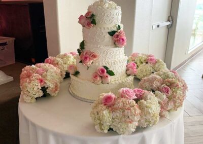 Wedding cake adorned with pink roses surrounded by floral arrangements; wedding flowers Orange County, modern floral design, fresh flowers delivery.