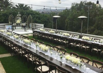 an elegantly arranged outdoor wedding reception area. The setting includes multiple long wooden tables adorned with white floral centerpieces and lined with white chairs that have dark brown frames.