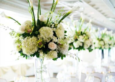 A series of tall, clear glass vases filled with an arrangement of white flowers, prominently featuring lilies and hydrangeas, accented with green foliage.