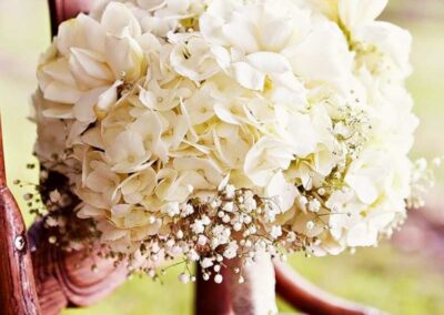 A bridal bouquet consisting of white flowers, including large blooms and smaller filler flowers, resting on the seat of a wooden chair with an ornate design. The bouquet is tied together with what appears to be a satin or silk ribbon. The background is softly blurred, highlighting the bouquet and giving the scene a romantic and elegant ambiance.