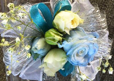 a floral arrangement consisting of blue and white flowers, with some smaller yellowish-white blooms interspersed throughout. The flowers are accented with a bright blue ribbon and delicate white netting or tulle.