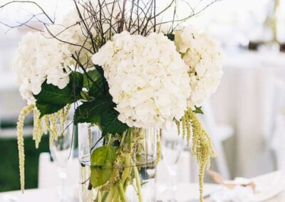 A floral arrangement used as a centerpiece, possibly for an event such as a wedding or formal gathering. The arrangement consists of white hydrangeas, with their large, rounded clusters of flowers providing a lush and full appearance. Accompanying the hydrangeas are cascading green amaranthus flowers, adding a sense of movement and contrast to the composition.