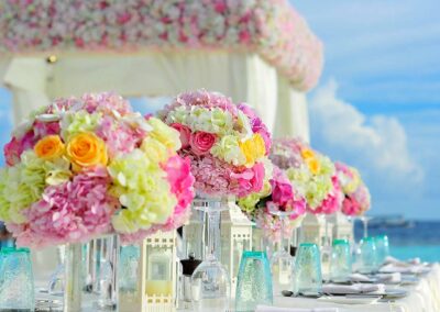 Vibrant floral arrangements with pink, yellow, and white flowers decorate an outdoor table setting by the seaside, under a clear blue sky.