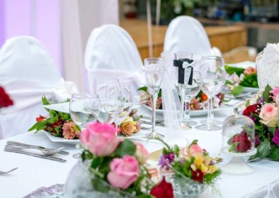 An elegant table setting, likely prepared for a special event such as a wedding or banquet. The table is adorned with crisp white linens and features a centerpiece composed of vibrant pink and red flowers interspersed with greenery.