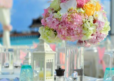 An outdoor table setting, highlighting a centerpiece composed of a lush bouquet of flowers with varying shades of pink, white, and yellow. The bouquet is arranged in a clear glass vase on top of the table.
