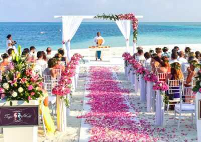 Beach wedding ceremony setup with white chairs and pink floral arrangements lining the aisle, ocean backdrop and a decorated archway.
