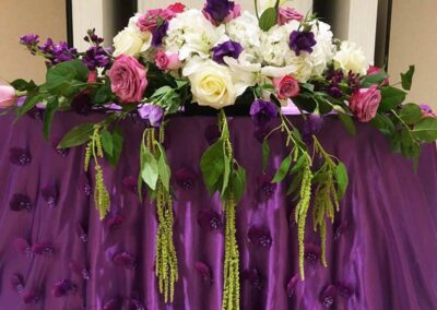 Elegant floral arrangement with white, pink, and purple flowers on a purple fabric-draped table; wedding and event flowers Orange County.