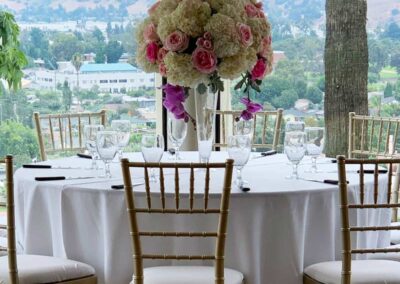 Round table with elegant gold chairs, featuring a tall centerpiece of white and pink floral arrangements, set for an event.